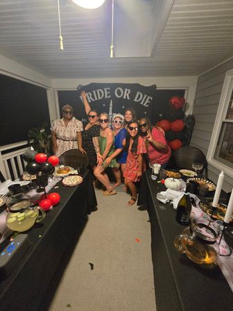 Seven friends posing on a covered porch at a bachelorette party in front of a 'BRIDE OR DIE' banner, surrounded by black-and-red balloons, long tables of snacks and drinks.