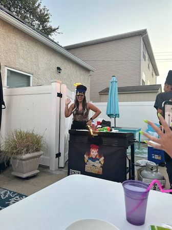 Backyard patio party scene with a woman in a novelty hat and sunglasses waving beside a grill with a small flame, white vinyl fence, turquoise umbrella, propane tank and a purple cup in the foreground.