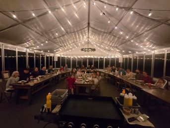 Community dinner in a clear-sided event tent at night — long wooden U-shaped tables, string lights overhead, guests dining around a central griddle and condiment station.