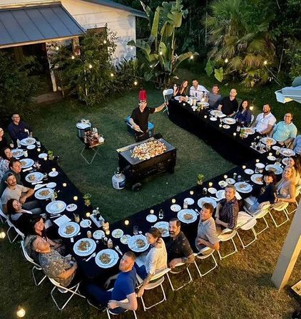 Aerial view of a backyard evening dinner party with U-shaped black-clothed tables, guests seated around plates and candles, a chef cooking on a central flat-top griddle, string lights and tropical plants creating a cozy garden atmosphere.