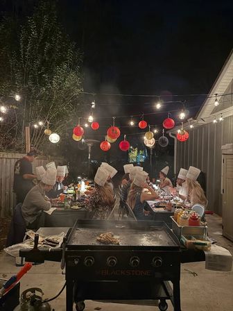 Backyard patio dinner at night with guests wearing chef hats seated at a long table under string lights and red paper lanterns, a steaming flat‑top griddle cooking in the foreground.