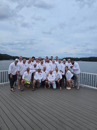 Large group of adults in matching white shirts posing on a lakeside deck by a calm lake under a cloudy sky — team retreat group photo.