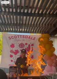 Chef performing a dramatic flambé at an outdoor Scottsdale event — tall orange flames under a pergola in front of a pink “Scottsdale Before the Veil” banner and yellow-pink balloon column, guest filming on phone.