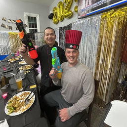 Two people at an indoor New Year celebration posing with colorful water guns — one wearing a tall red novelty hat — metallic fringe backdrop, gold "2023" balloons and a table with plates and drinks.