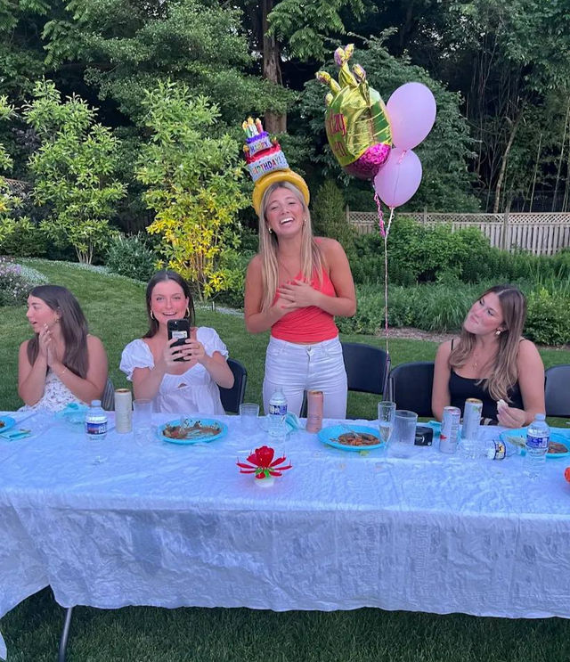 Four friends at an outdoor backyard birthday party seated at a long white table with plates and drinks; smiling celebrant in a pink top wearing a colorful cake-shaped birthday hat and holding pink balloons in a green garden setting