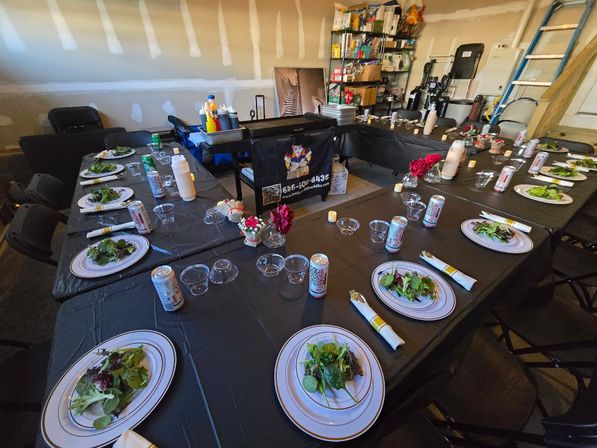 Casual garage dinner setup in U-shaped layout with black tablecloths, plates of mixed green salads, canned drinks, clear plastic cups, small floral centerpieces, condiment station and folding chairs.