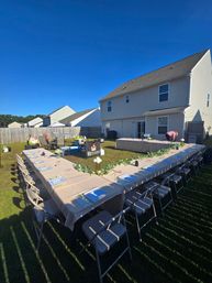 U-shaped banquet table with beige tablecloths, place settings, folding chairs and green garland set for an outdoor garden party in a sunny suburban backyard by a two-story house.