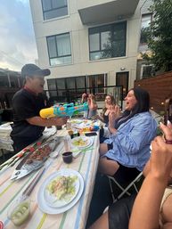 Group of friends laughing at an urban backyard patio dinner in front of a modern apartment as a guest playfully aims a colorful water gun across a table set with salads and skewers.
