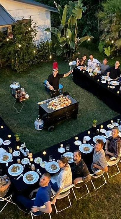 Aerial view of a backyard evening dinner party with U-shaped black-clothed tables, guests seated around plates and candles, a chef cooking on a central flat-top griddle, string lights and tropical plants creating a cozy garden atmosphere.