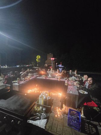 Nighttime backyard patio party on a paved patio — guests around a U-shaped table lit by string lights and candles, raising glasses; grill and condiment bottles in the foreground, patio heater and gold '2' balloon in the background.