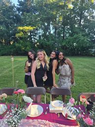Four smiling friends posing behind chairs at a backyard summer dinner party with a pink floral tablescape, drinks, tiki torch, and tree-lined lawn