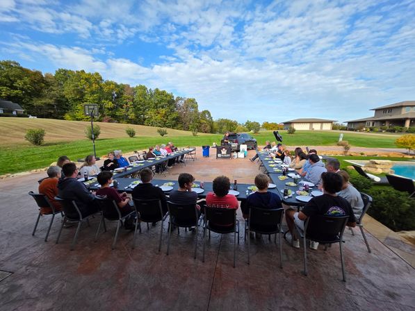 Fun suburban backyard dinner on a spacious patio: U-shaped long tables with family and friends seated near a pool and wide lawn, autumn trees and a blue sky with scattered clouds.