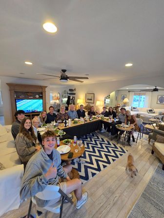 Large multigenerational group gathered for dinner around a long table in a bright coastal-style living room — salads, drinks, a TV showing football, ceiling fan, chevron rug, and a small dog crossing the hardwood floor.