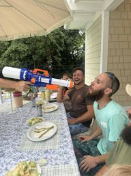 Friends at a backyard patio table laughing as someone sprays a large blue-and-orange water blaster into a bearded man's mouth under a patio umbrella during a summer outdoor meal.