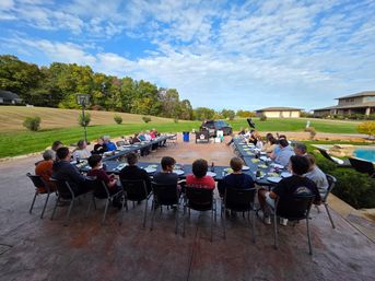 Fun suburban backyard dinner on a spacious patio: U-shaped long tables with family and friends seated near a pool and wide lawn, autumn trees and a blue sky with scattered clouds.