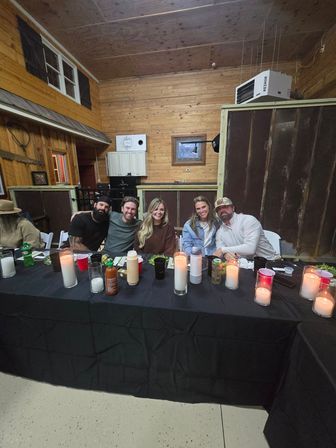 Five people smiling and leaning together at a long black-tablecloth table in a rustic wood-paneled barn, surrounded by lit pillar candles, drinks, and condiments for a cozy indoor farmhouse gathering.