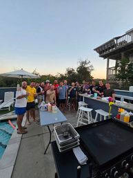 Poolside sunset gathering: a group of people raising drinks on a backyard patio with an outdoor griddle, bar stools, umbrella and a raised beach-style deck.