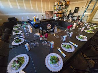 Casual garage dinner setup in U-shaped layout with black tablecloths, plates of mixed green salads, canned drinks, clear plastic cups, small floral centerpieces, condiment station and folding chairs.