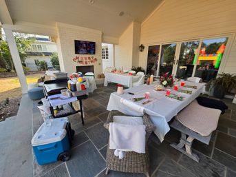 Festive backyard covered patio set for a Christmas party with white‑clothed tables, red cups and wrapped place settings, buffet cart and cooler, Merry Christmas banner on the fireplace, and a colorful inflatable bounce house visible through sliding doors