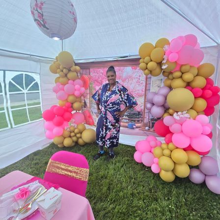 Woman in a navy floral kimono posing in a backyard party tent with large pink and gold balloon arches, cherry blossom backdrop, hanging paper lantern, and pink table setting on green grass.
