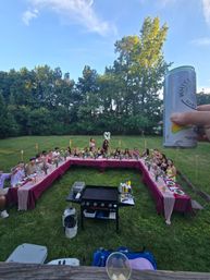 Backyard summer dinner party with a U-shaped table dressed in burgundy tablecloths, guests seated and toasting, tiki torches and floral centerpieces on a grassy lawn, tall trees under a blue sky, hand holding a canned drink in the foreground