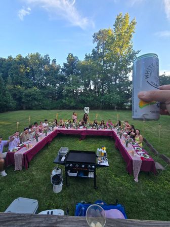 Backyard summer dinner party with a U-shaped table dressed in burgundy tablecloths, guests seated and toasting, tiki torches and floral centerpieces on a grassy lawn, tall trees under a blue sky, hand holding a canned drink in the foreground