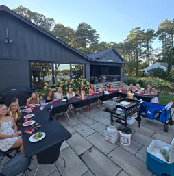 Group of friends enjoying an outdoor dinner on a modern lakefront home's backyard patio — long tables, grill, red cups, and summer trees.