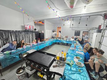 Indoor garage party with a large group seated around U-shaped tables covered in shiny blue tablecloths, colorful string lights overhead, a flat-top griddle and condiments in the foreground, and plates of food as kids and adults smile.