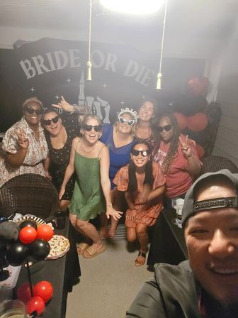 Smiling group of eight friends on a covered patio at night celebrating a bachelorette party; bride-to-be wearing 'BRIDE' heart sunglasses, black-and-red balloons and a 'BRIDE OR DIE' banner in the background.