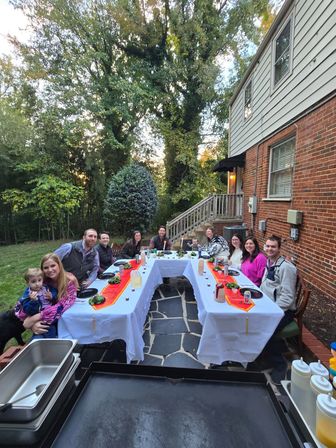 Smiling group gathered around a U-shaped table with white tablecloths and orange runners for an outdoor dinner on a stone patio beside a brick house, surrounded by tall trees in a leafy backyard at dusk.