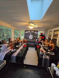 Group of friends toasting at a 'Bride or Die' bachelorette party on a screened-in porch in Richmond, long black-draped tables with candles and red-and-black balloons beneath a skylight and ceiling fan.
