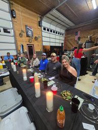 People enjoying a casual rustic dinner in a wood-paneled garage/workshop — long black table with lit pillar candles, salads, drinks, mounted deer heads and an American flag.