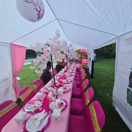 Outdoor pink-and-gold party tent on a lawn with a long banquet table set for celebration — vibrant pink tablecloths and chair covers with gold sashes, faux cherry blossom centerpieces, paper lanterns overhead, gold cutlery place settings, and a pink-gold balloon garland at the far end.