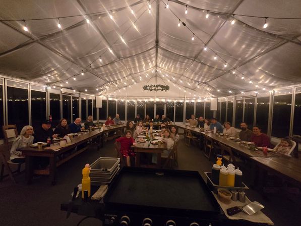 Community dinner in a clear-sided event tent at night — long wooden U-shaped tables, string lights overhead, guests dining around a central griddle and condiment station.