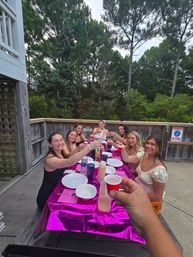 Group of friends toasting with small red cups around a shiny pink-covered table on a wooden outdoor deck surrounded by tall pine trees.