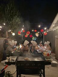 Backyard patio dinner at night with guests wearing chef hats seated at a long table under string lights and red paper lanterns, a steaming flat‑top griddle cooking in the foreground.
