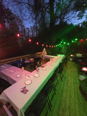 Festive backyard wooden deck at dusk with a U-shaped table set for dinner, red and green string lights and a small lit Christmas tree.