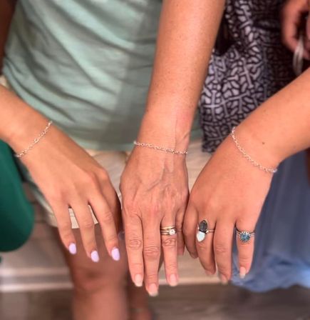 Close-up of three women's hands wearing matching delicate silver chain bracelets and assorted rings with gemstone accents, one with light pastel nail polish and a casual summer clothing backdrop.