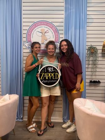 Three smiling people posing inside a pastel-themed boutique, holding a round “I got ZAPPED” sign in front of blue curtains and a slatwall display with a circular logo, pink armchairs visible.
