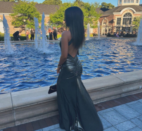 Back view of a person in a sleek black backless evening gown posing by a blue fountain in an outdoor plaza with an arched building and guests in the background.
