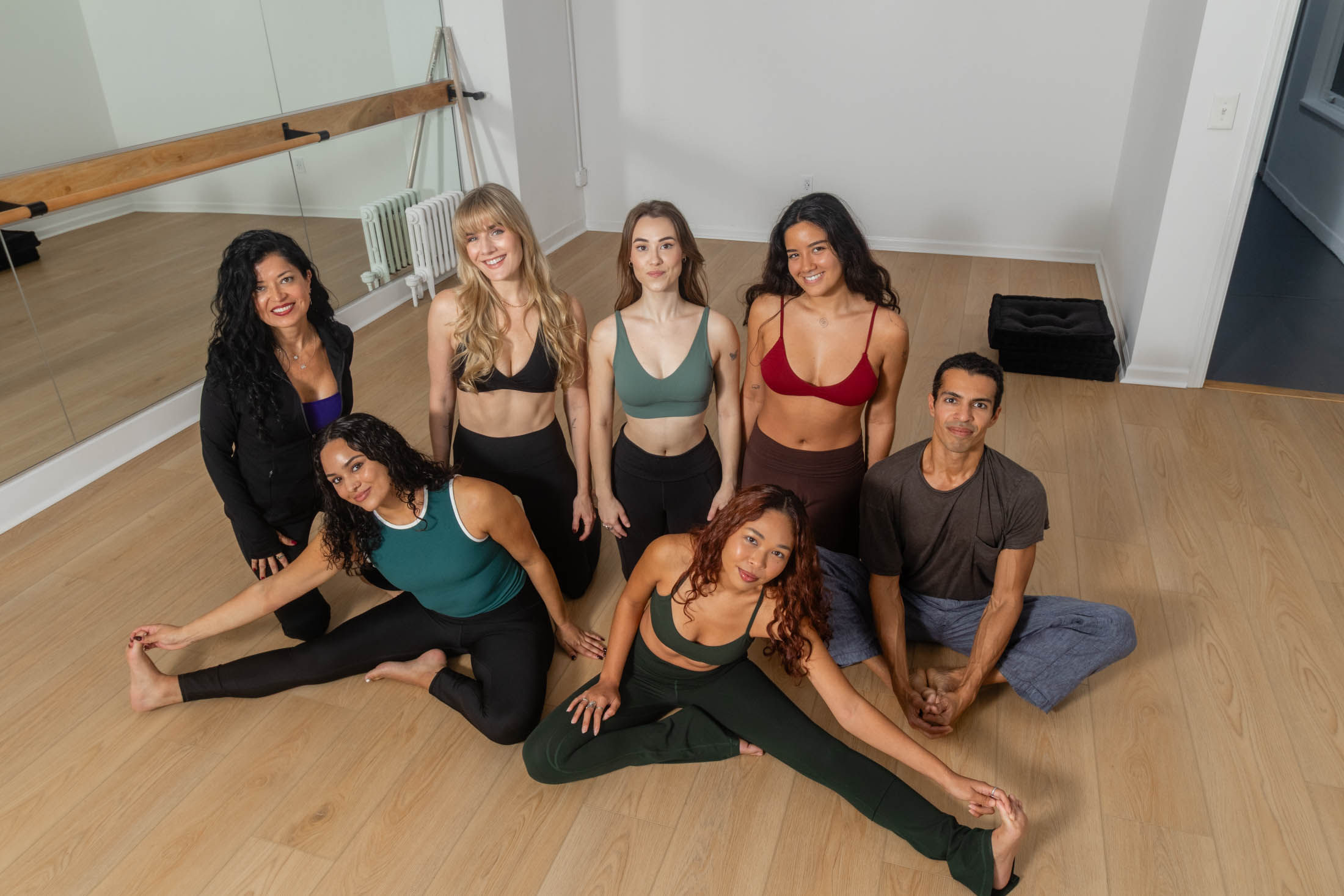 Group of eight adults in colorful activewear smiling and stretching on a wooden floor in a bright indoor yoga studio with mirrors and a barre — group fitness/yoga class setting.
