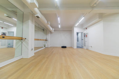 Spacious modern dance studio with floor-to-ceiling mirrors, wooden ballet barre, light oak floors, white walls and bright overhead lighting.