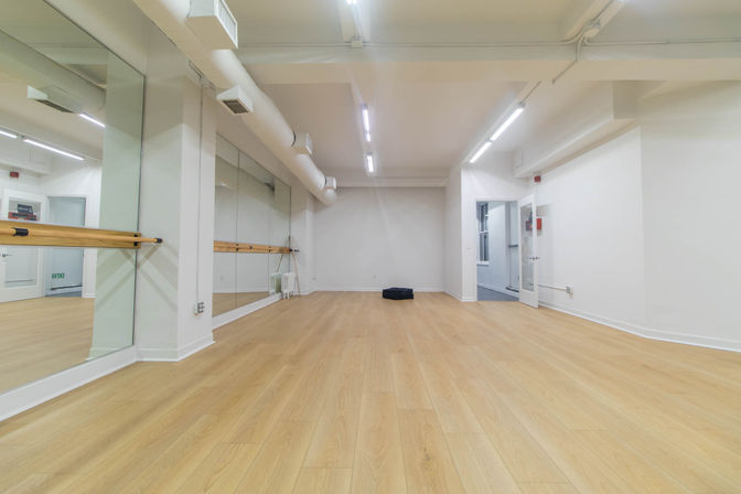 Spacious modern dance studio with floor-to-ceiling mirrors, wooden ballet barre, light oak floors, white walls and bright overhead lighting.