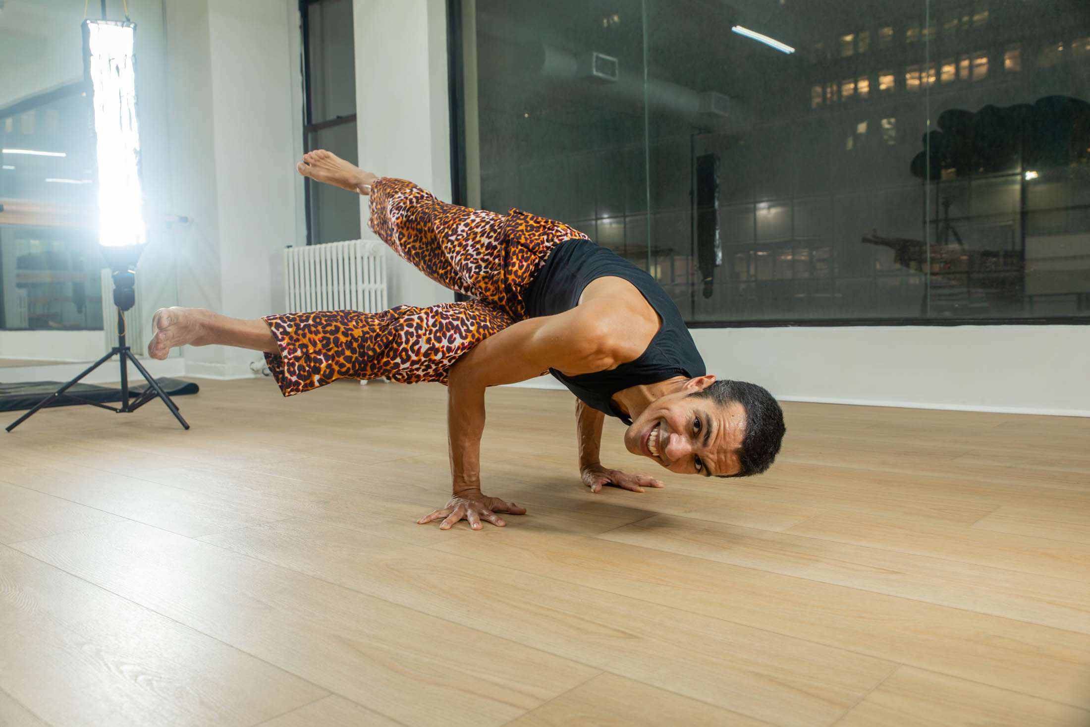 Yogi smiling in an advanced arm-balance pose on hands in an urban studio, wearing leopard-print pants and a black tank top on a light wood floor with studio lights and city windows behind.