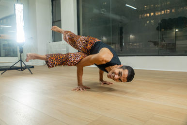 Yogi smiling in an advanced arm-balance pose on hands in an urban studio, wearing leopard-print pants and a black tank top on a light wood floor with studio lights and city windows behind.