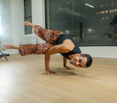 Yogi smiling in an advanced arm-balance pose on hands in an urban studio, wearing leopard-print pants and a black tank top on a light wood floor with studio lights and city windows behind.