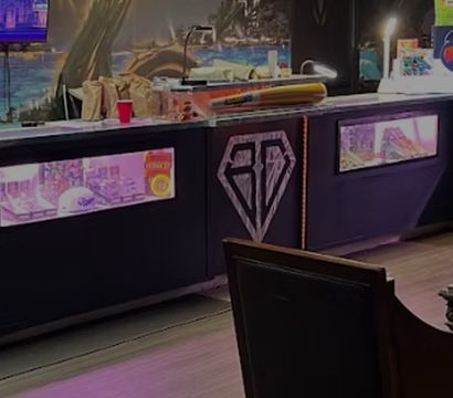 Cozy neon-lit indoor lounge with a long glass display counter beneath a colorful mural, plush armchairs around a carved wooden coffee table, ambient lamps and modern decor
