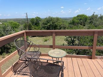 Two wrought-iron patio chairs and a small mosaic table on a wooden deck overlooking oak‑covered rolling hills under a clear blue sky.