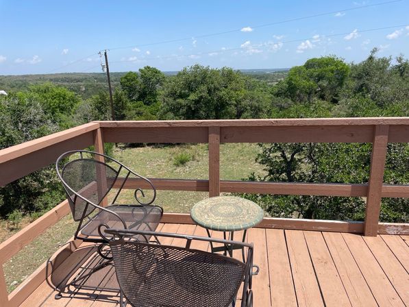 Two wrought-iron patio chairs and a small mosaic table on a wooden deck overlooking oak‑covered rolling hills under a clear blue sky.