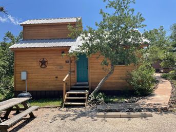 Rustic tiny wooden cabin with metal roof and teal front door, front steps and star wall ornament, picnic table and grill on gravel, shaded by a leaning oak and a winding stone path.